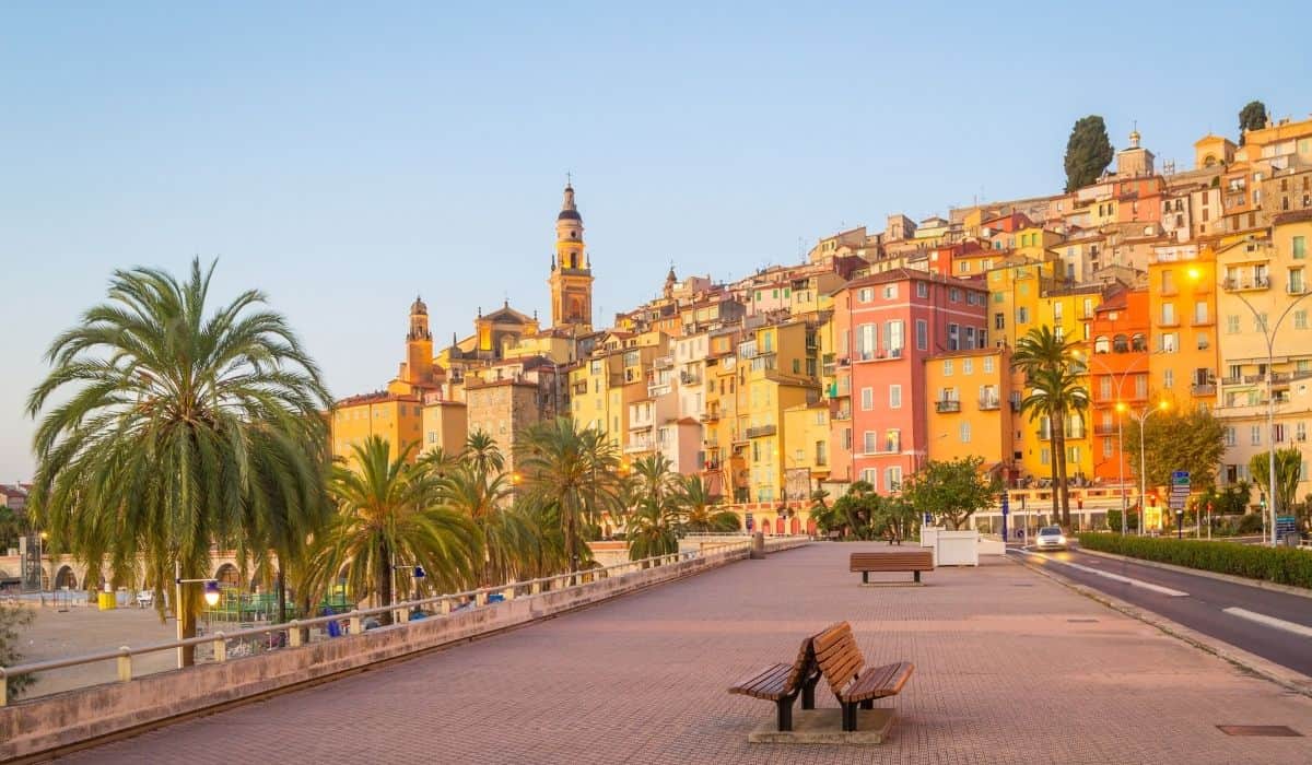 Promenade along the water with colorful buildings and palm trees in Menton, France.